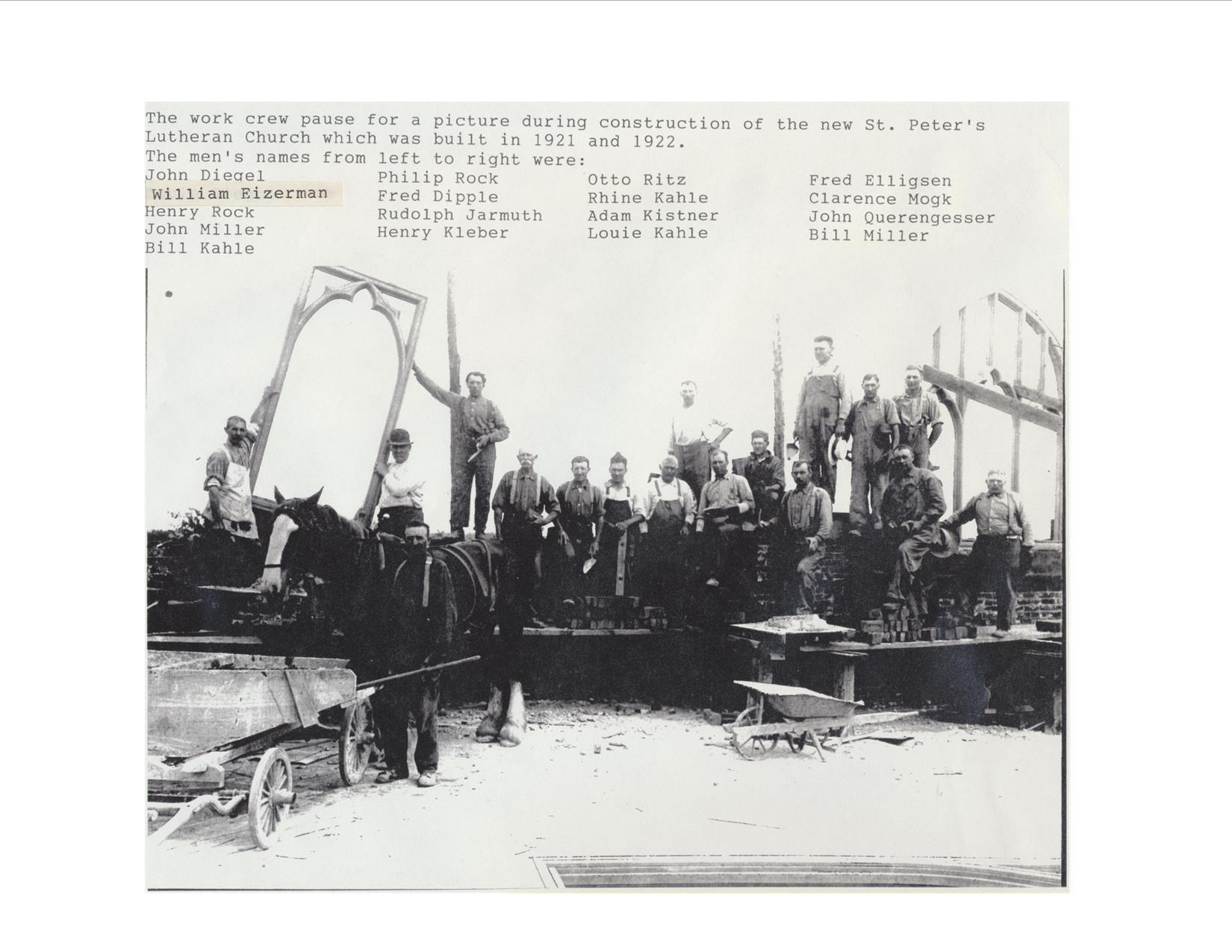 17 men posed for picture at construction site with a horse, tools and frames of what will be stained glass windows